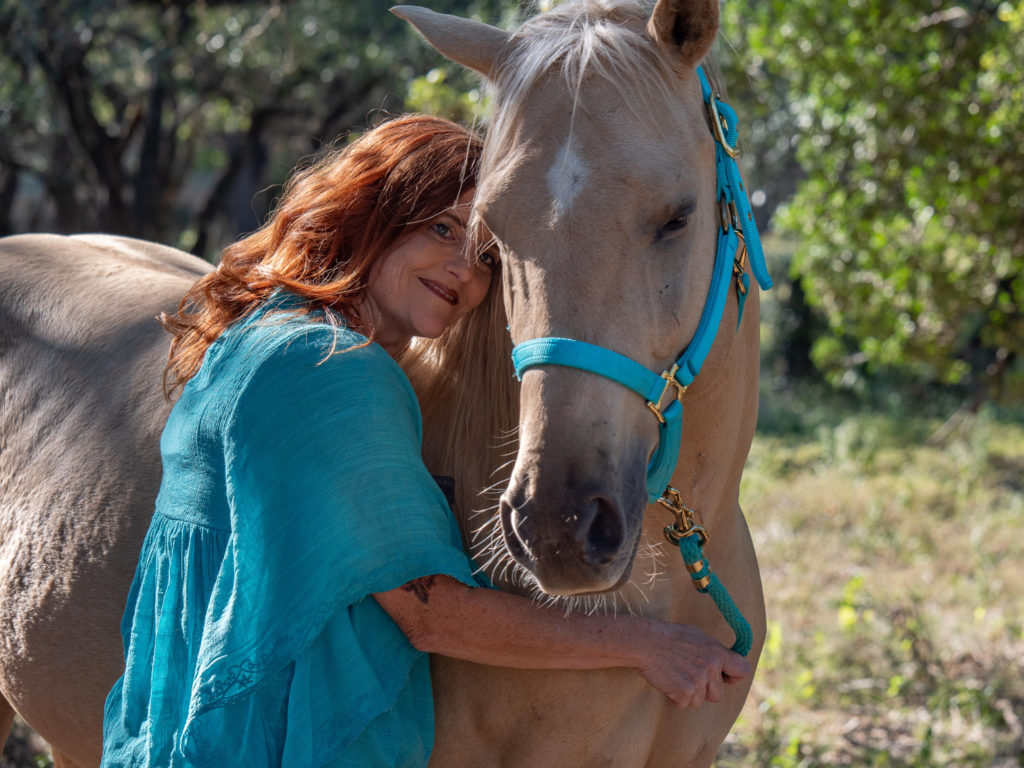 Julie Bradshaw, woman with long red hair, leaning on neck of Palomino horse with turquoise halter