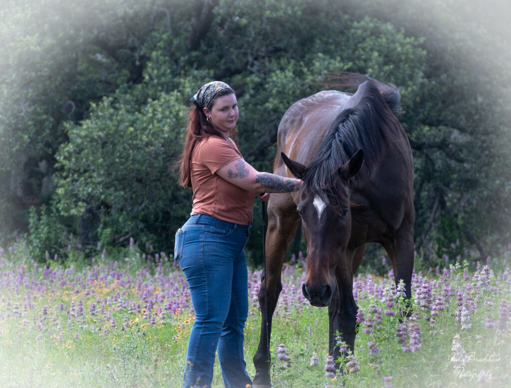 Young woman with long brown hair, colorful bandana on her head, orange shirt, and blue jeans pets a dark bay horse on the shoulder during a Solitude with Horses session.